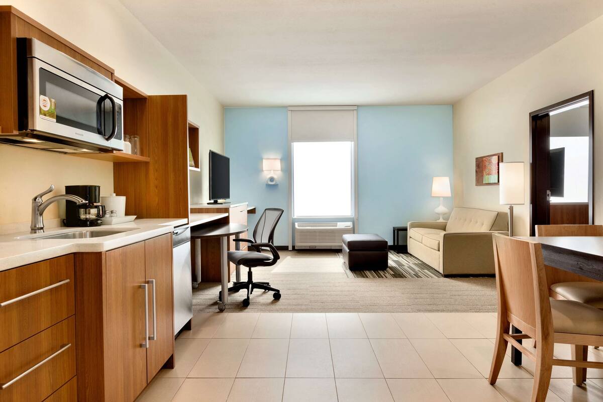 A modern hotel room with a kitchenette, a dining area, a sofa, and a TV.