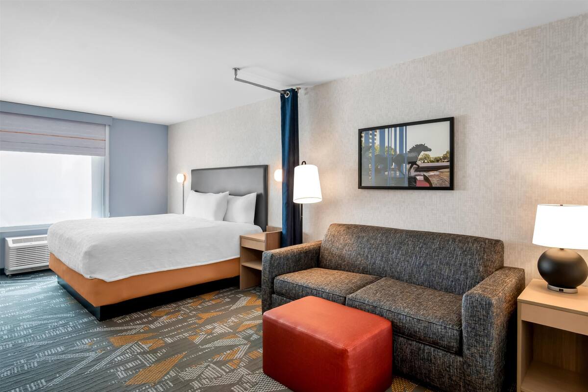 Een hotelkamer met een bed, een bank, een rode poef, een nachtkastje, een lamp en een ingelijst foto aan de muur.