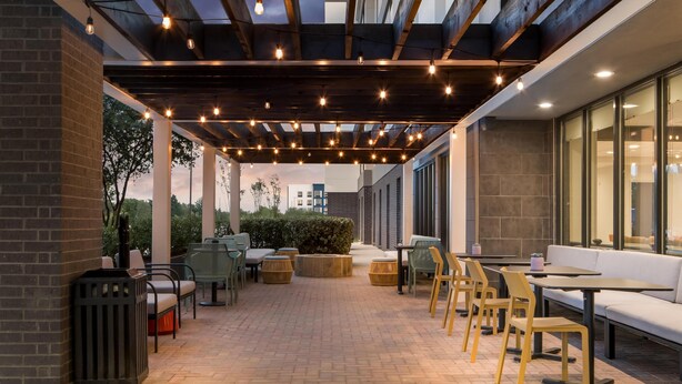 An outdoor patio area with seating arrangements, a pergola with string lights, and a view of the surrounding buildings. - Home2 Suites by Hilton Irving / DFW Airport North (Irving)