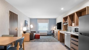 A compact hotel room with a kitchenette, a dining area, a seating area with a sofa and ottoman, a large window with blinds, and a flat-screen TV. - Home2 Suites by Hilton Irving / DFW Airport North (Irving)
