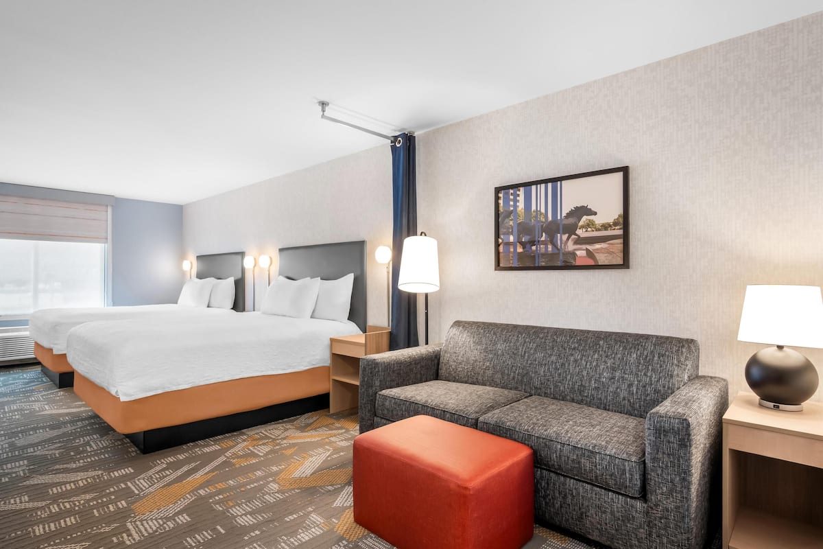 Hotelkamer met twee bedden, een bank, een rode poef, een lamp, een schilderij aan de muur en een raam met jaloezieën.