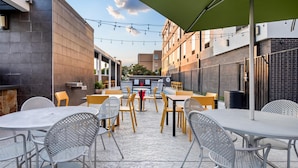 An outdoor patio area with round tables and chairs, string lights, and a green umbrella. - Home2 Suites by Hilton Irving / DFW Airport North (Irving)
