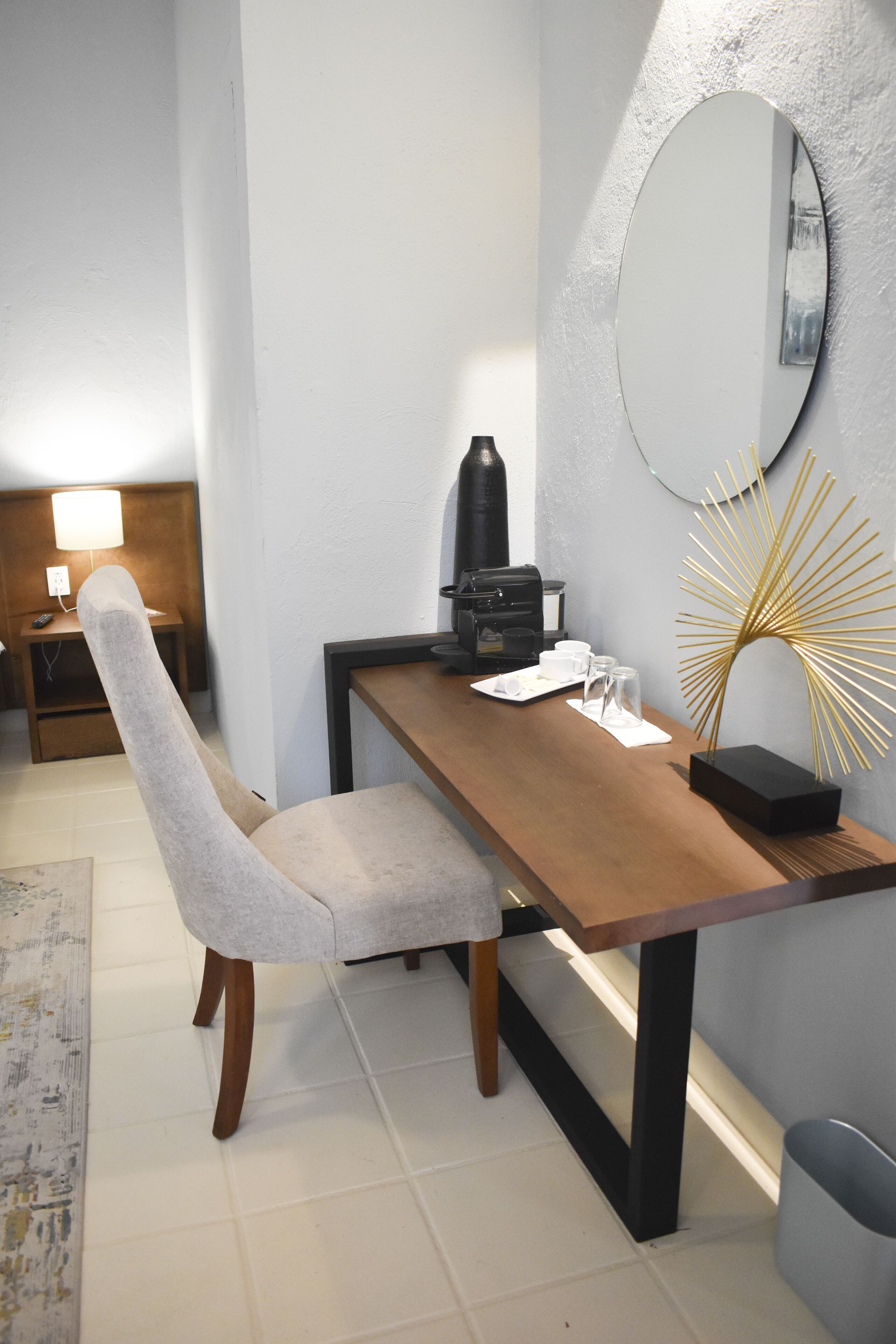 Une chambre d’hôtel moderne avec un bureau en bois, un fauteuil rembourré de couleur grise, une machine à café, un miroir et une plante décorative.