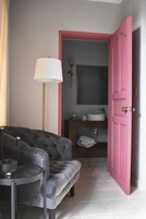 Una habitación con un sofá gris, una lámpara de pie, una puerta rosa y un baño visible a través de una puerta abierta.