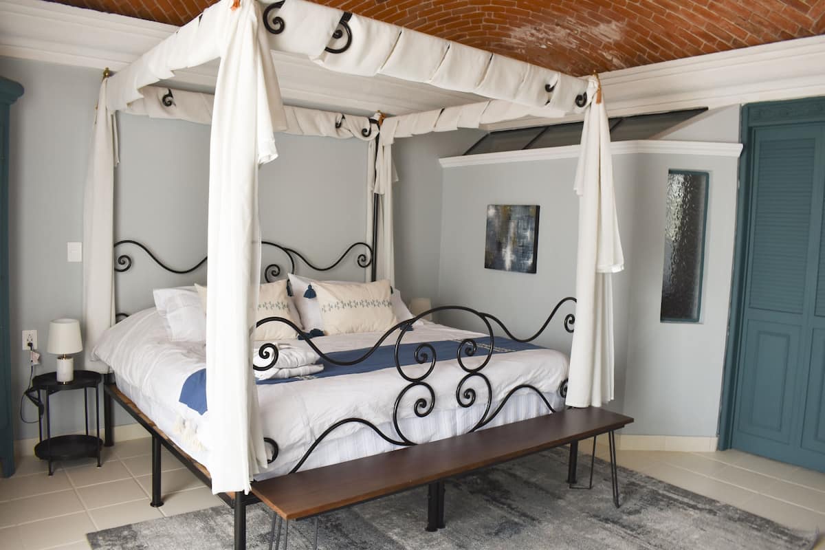 A four-poster canopy bed with a bench in front, a bedside table with a lamp, and a blue door in the background.