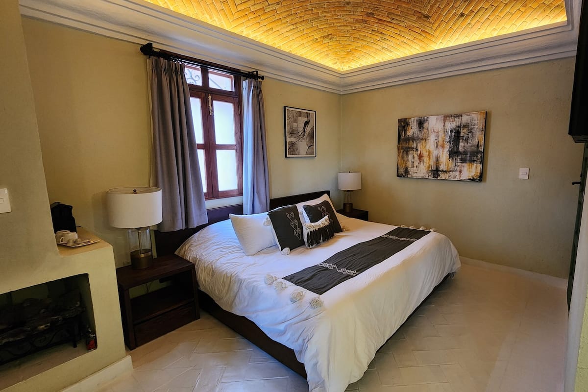 A hotel room with a bed, a nightstand, a lamp, a painting, and a fireplace.