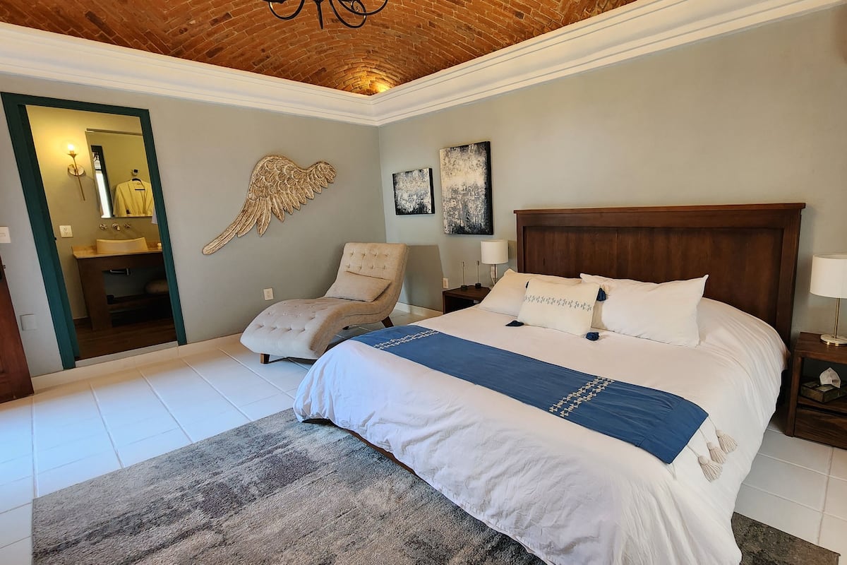 A bedroom with a large bed, a wooden headboard, a nightstand with a lamp, a chair, a mirror, and artwork on the walls.