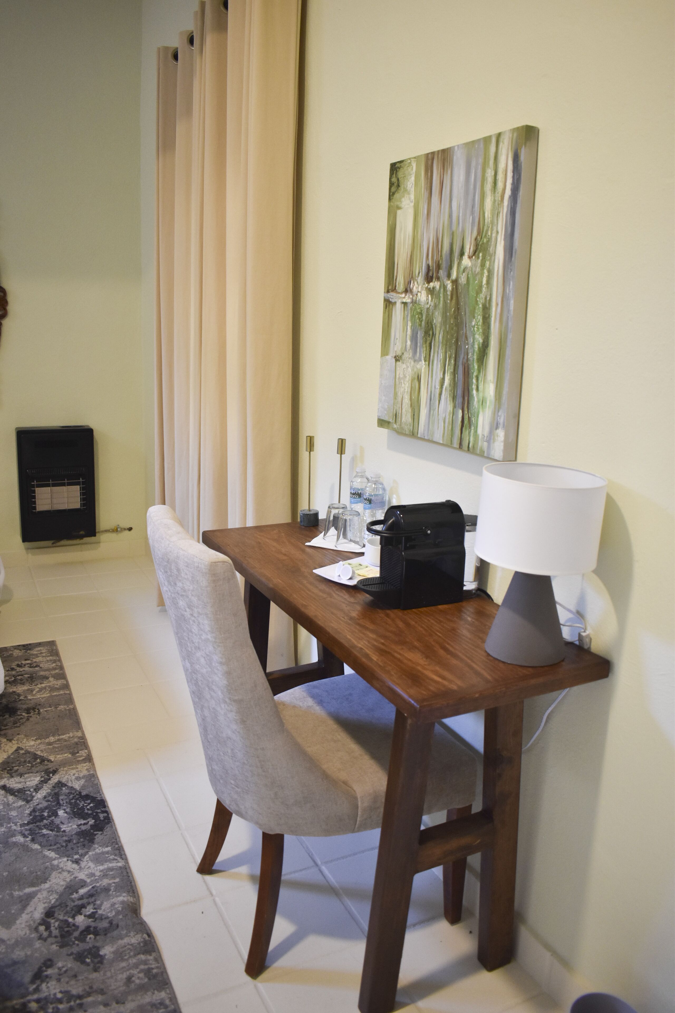 A hotel room with a wooden desk, a chair, a lamp, a painting on the wall, and a radiator.