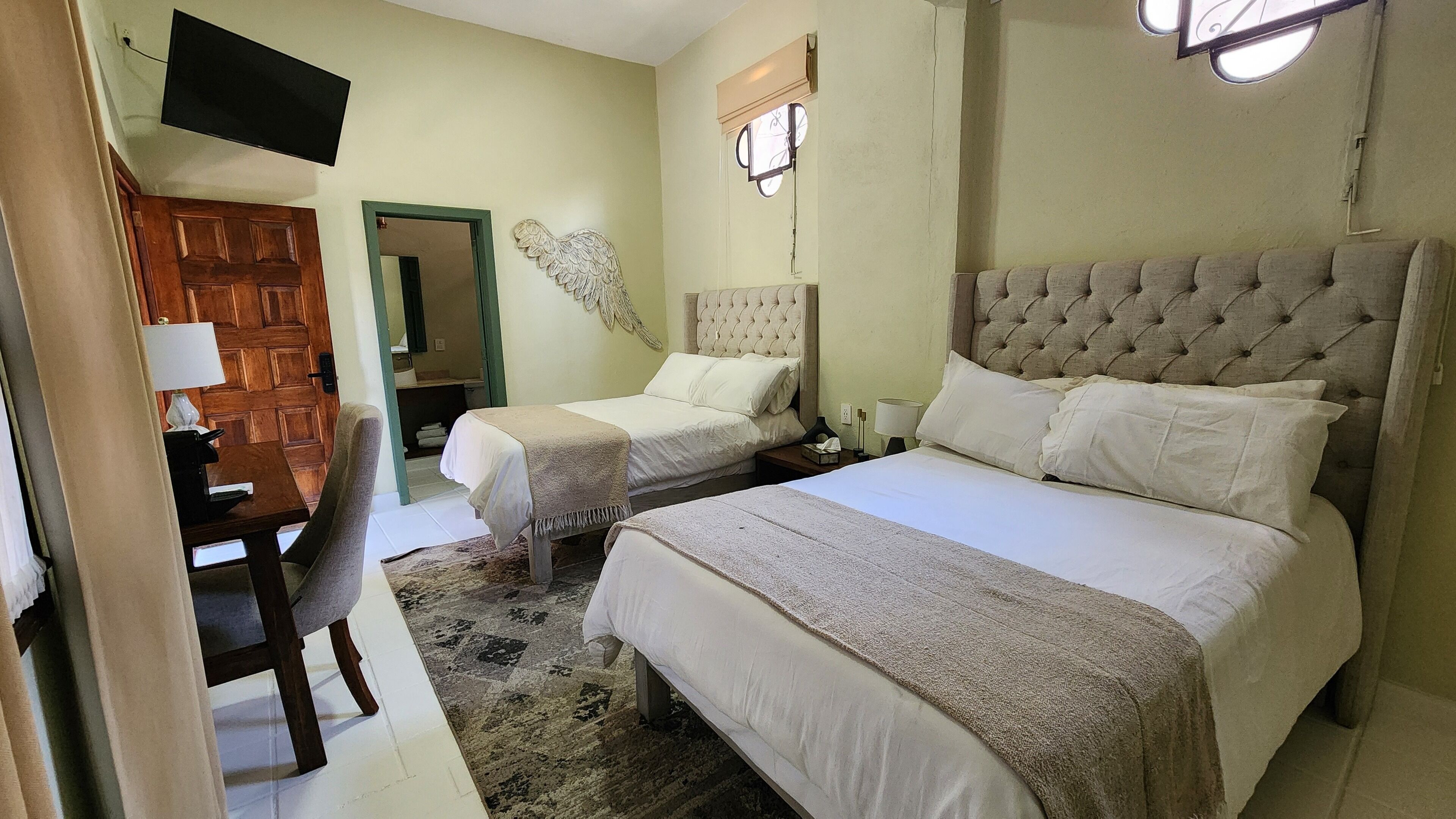 A hotel room with two beds, a desk, a chair, a TV, and a window with a clock.