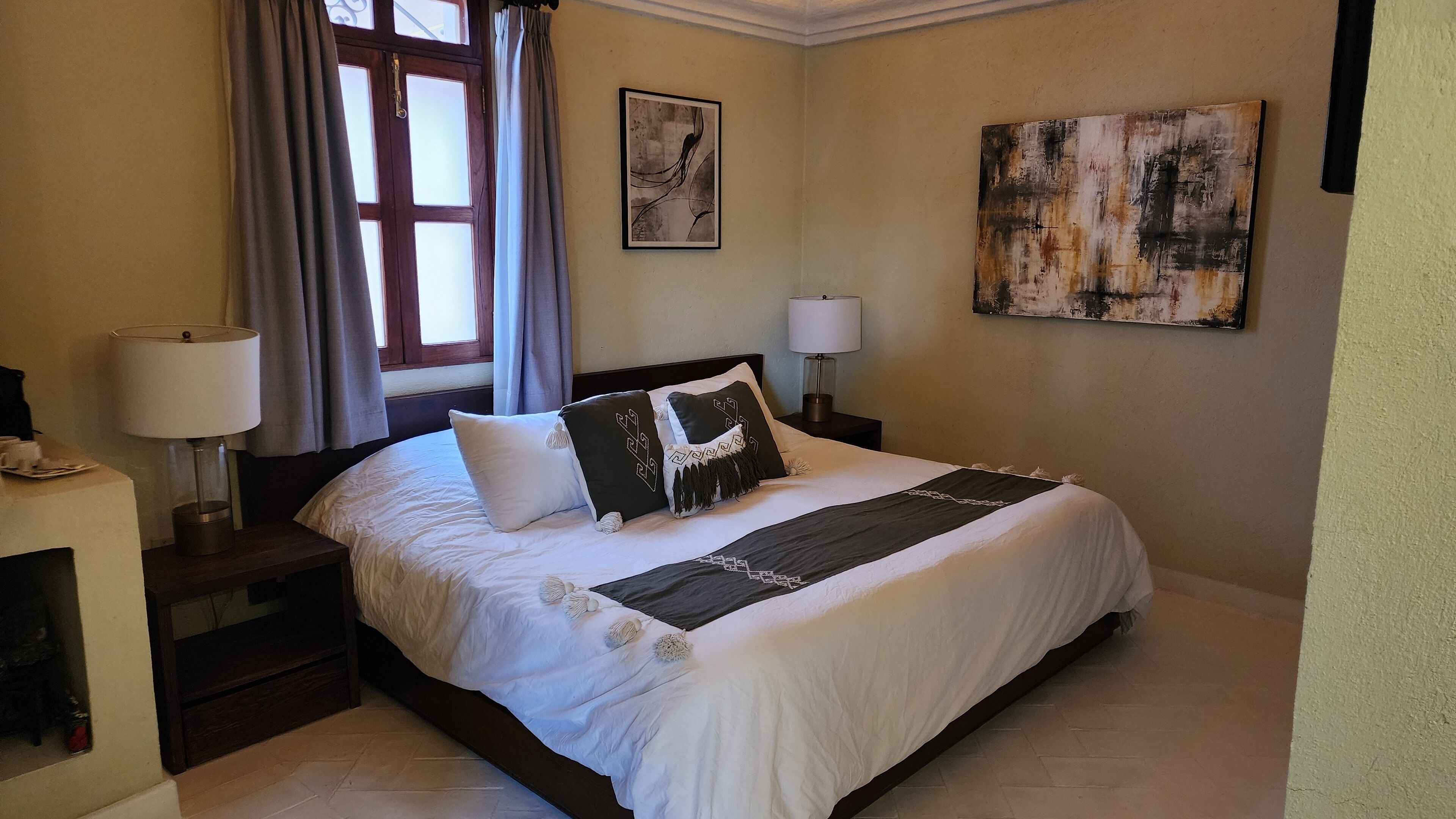 A neatly made bed with a white and dark striped bedspread, two bedside lamps, a window with curtains, and two wall art pieces.