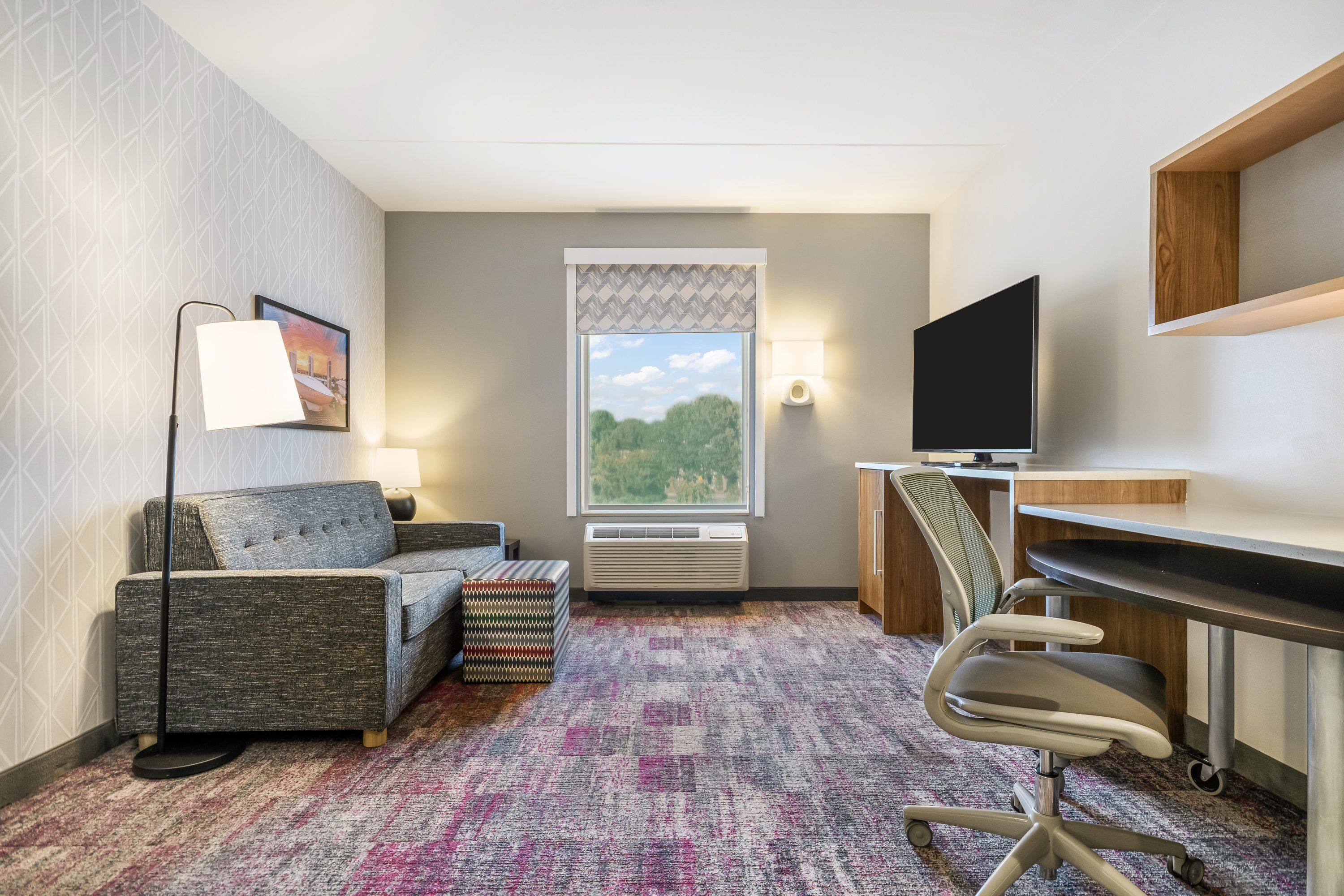 A hotel room with a sofa, a desk, a chair, a television, a window with a view, and a patterned carpet.