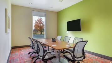 A conference room with a long table, six chairs, a flat-screen TV on the wall, and a window with a view of trees.
