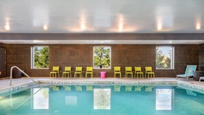 A swimming pool with clear water, surrounded by yellow chairs and a pink table, with windows offering views of trees. - Home2 Suites by Hilton Albany Wolf Rd (Albany)