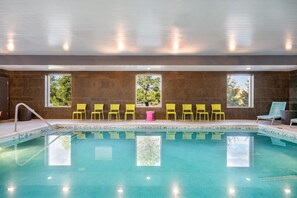 A swimming pool with clear water, surrounded by yellow chairs and a pink table, with windows offering views of trees. - Home2 Suites by Hilton Albany Wolf Rd (Albany)