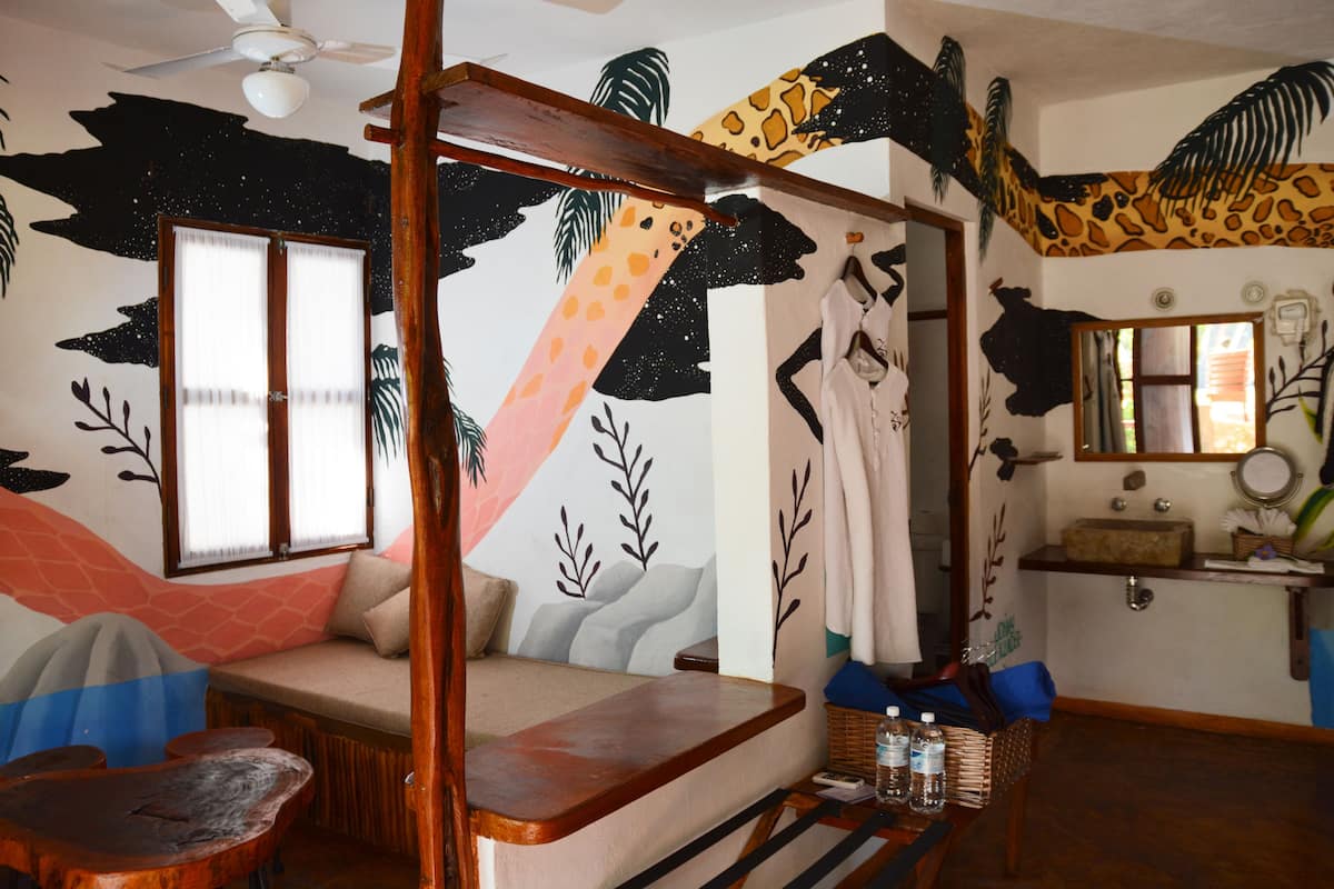 Une chambre avec un lit à baldaquin, un banc en bois, une petite table avec des bouteilles et un miroir au-dessus du lavabo.