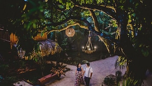 A couple standing in a garden with a thatched structure and hanging lights.