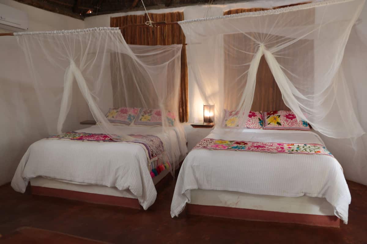 Deux lits avec du linge de lit blanc et des motifs floraux colorés, un plancher en bois et un climatiseur blanc fixé au mur.