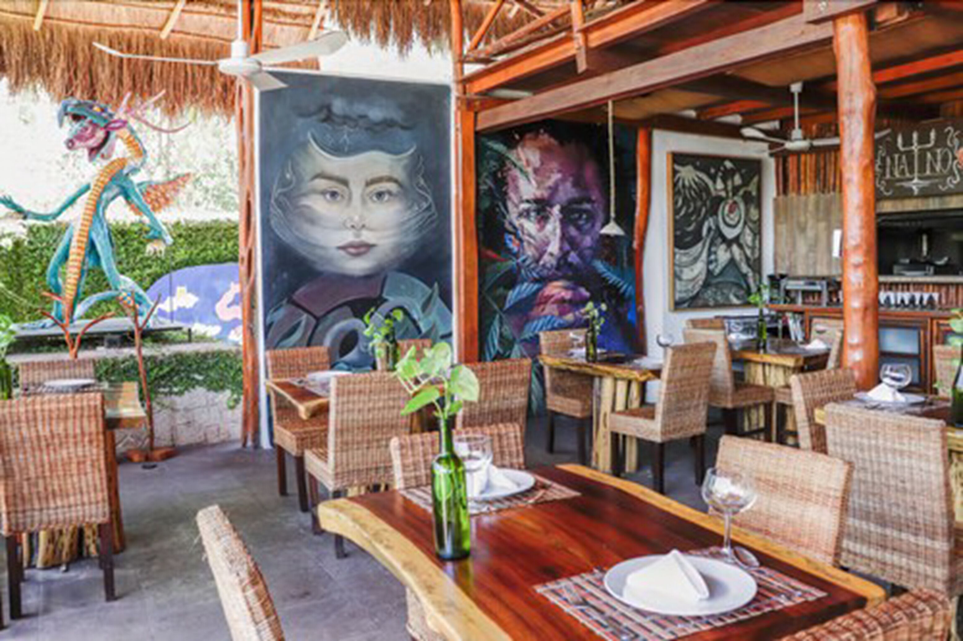 Un área de comedor al aire libre con mobiliario de mimbre, una mesa de madera y un mural en la pared.
