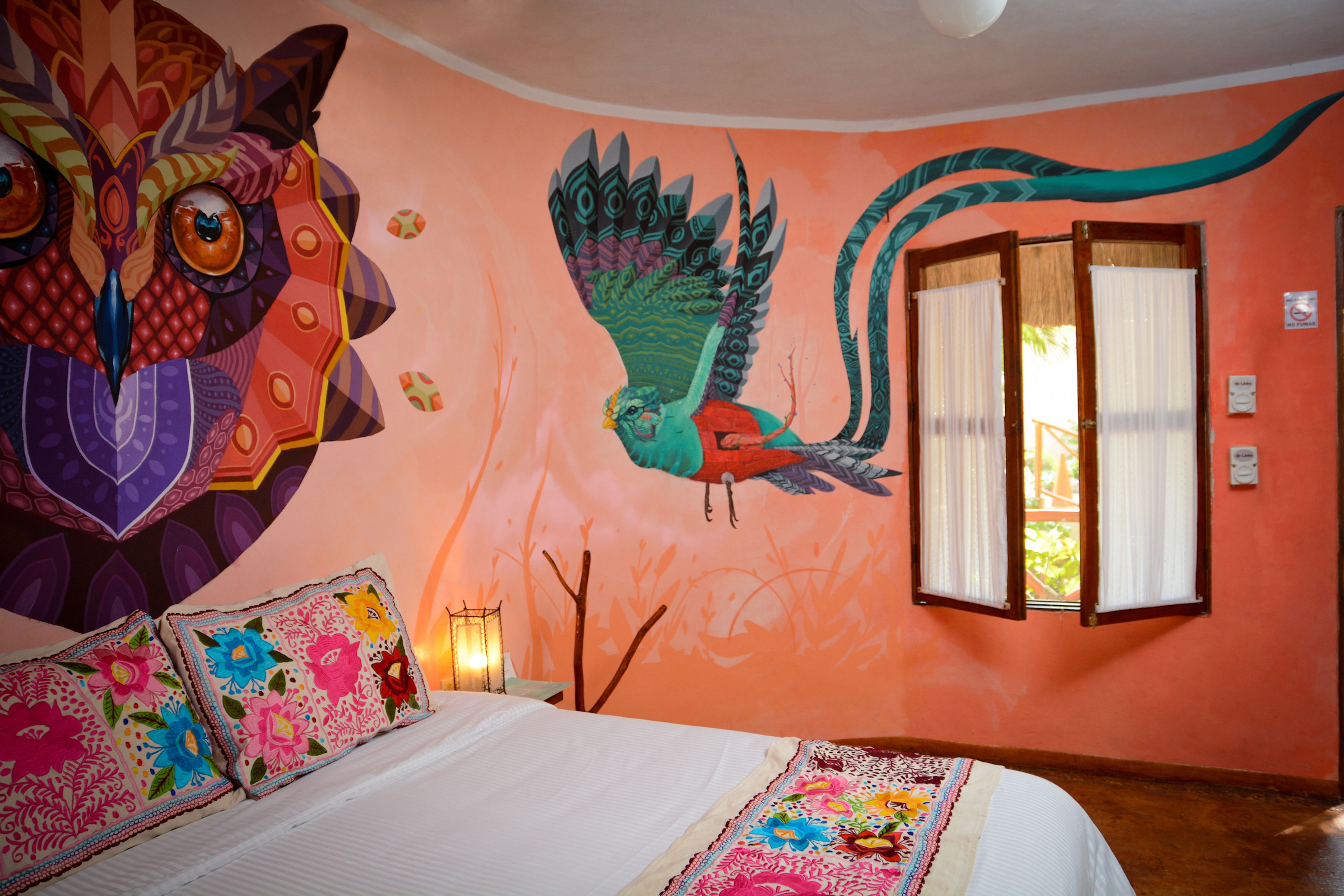 A room with a large mural of a bird, a bed with colorful pillows, and a window with curtains.