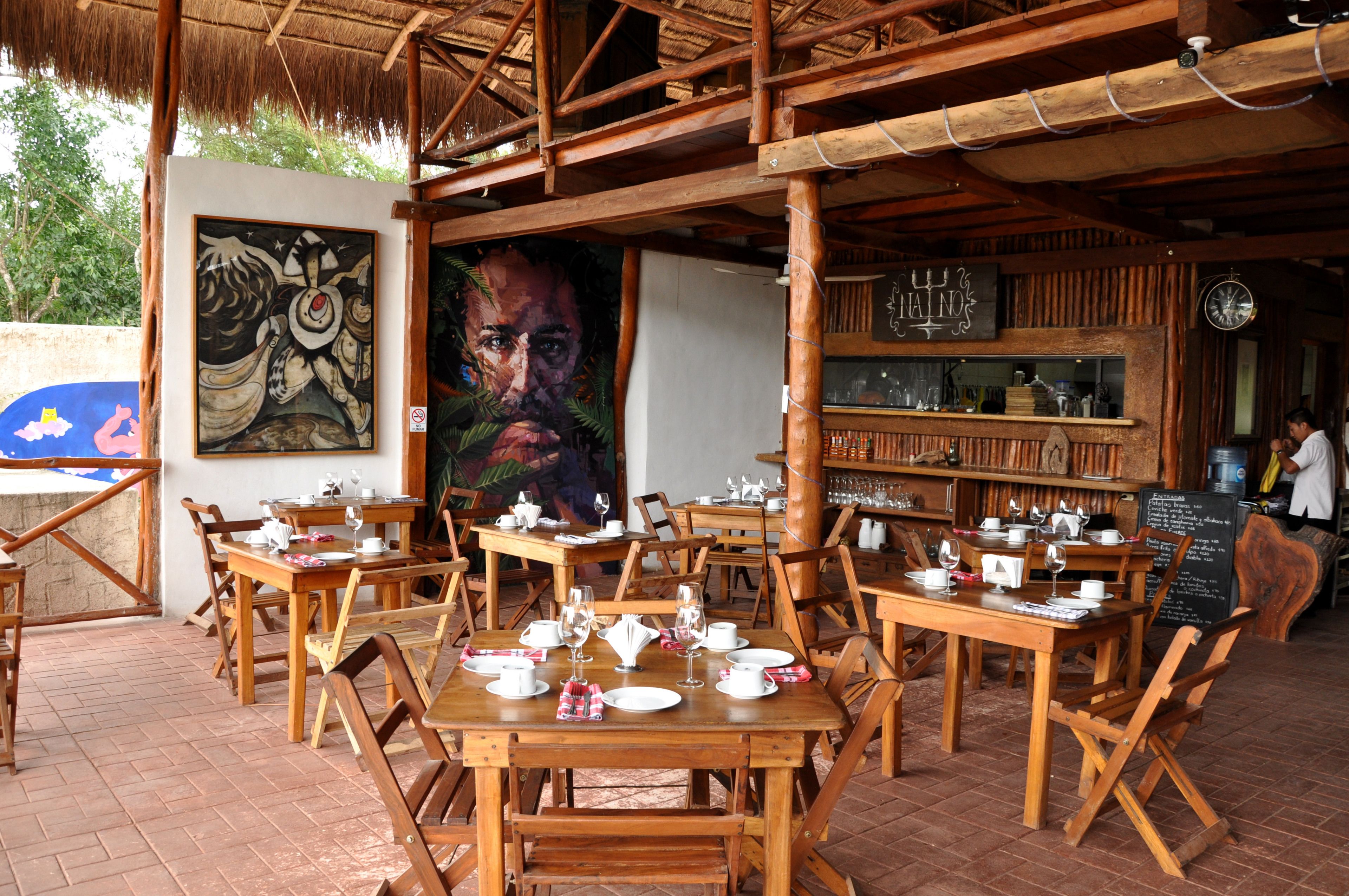 Un café en plein air avec des tables et des chaises en bois, une fresque murale et un coin bar avec des étagères remplies de bouteilles.