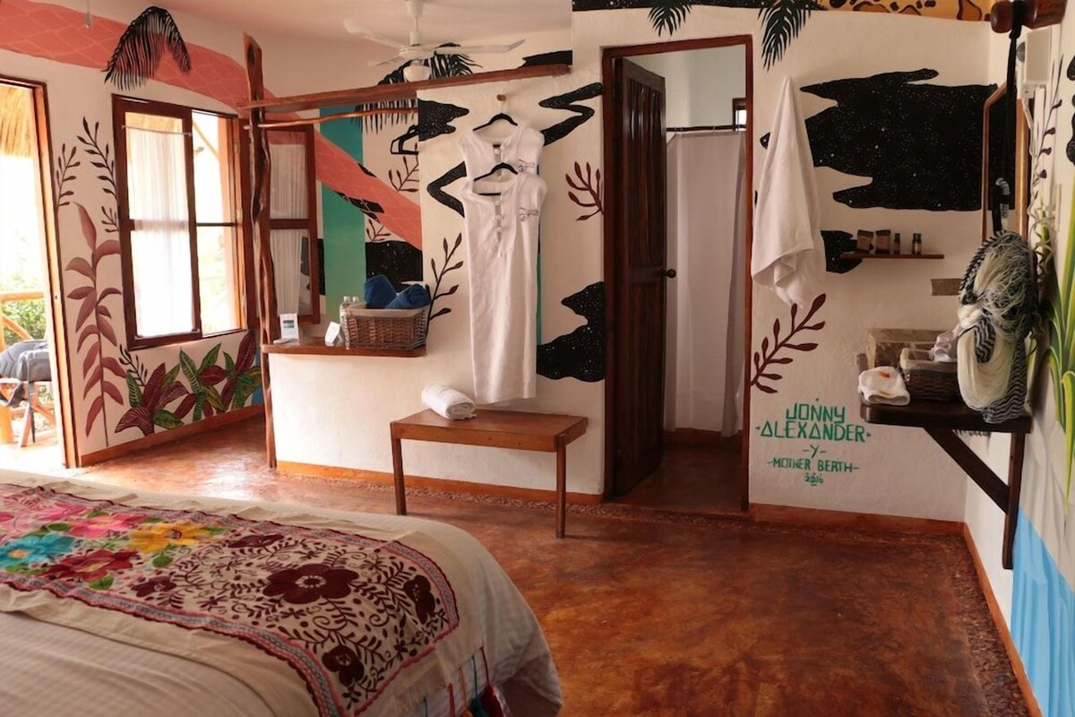Een kamer met een bed, een bankje, een spiegel en een muur met een naam en titel.