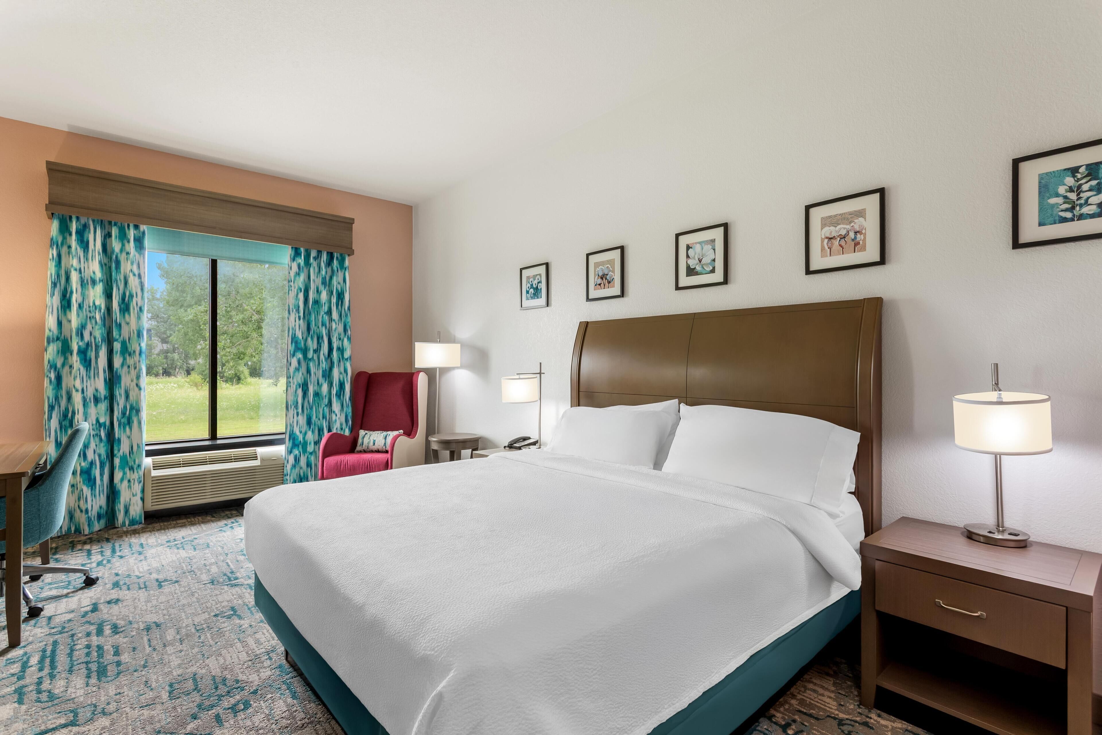 Habitación de hotel con una cama grande, un escritorio, una silla, una mesita de noche con lámpara y una ventana con cortinas.