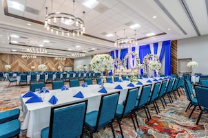 Un salón de banquetes con filas de mesas, sillas azules y centros de mesa florales.