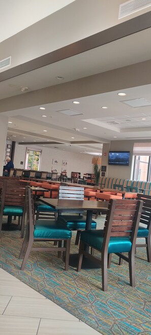A spacious dining area with multiple tables and chairs, a flat-screen TV mounted on the wall, and a patterned carpet. - Hilton Garden Inn Bolingbrook I-55 (Bolingbrook)