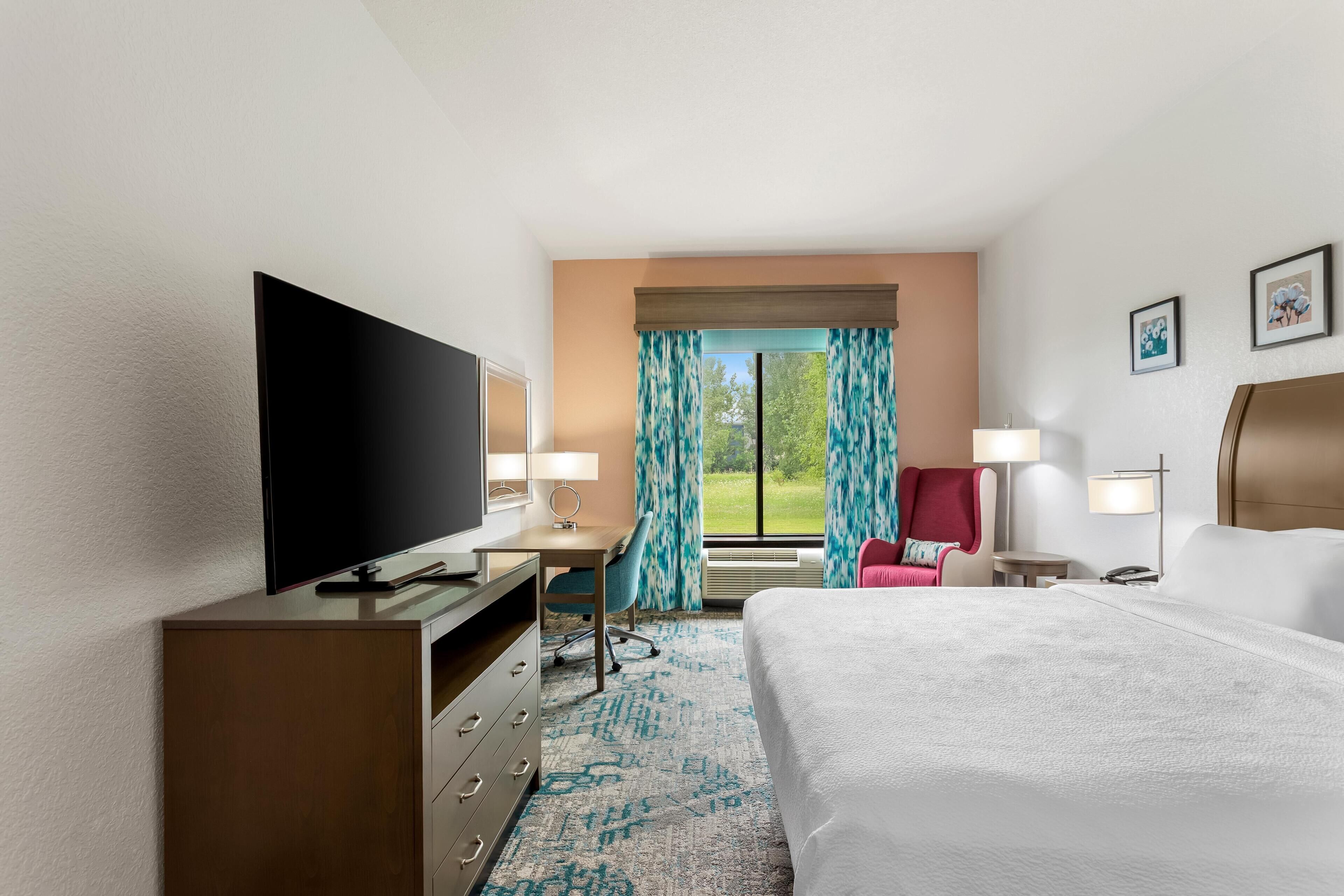 Een hotelkamer met een bed, een bureau met een flatscreen televisie, een stoel, een rode fauteuil, een lamp en een raam met gordijnen.