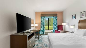Een hotelkamer met een bed, een bureau met een flatscreen televisie, een stoel, een rode fauteuil, een lamp en een raam met gordijnen.