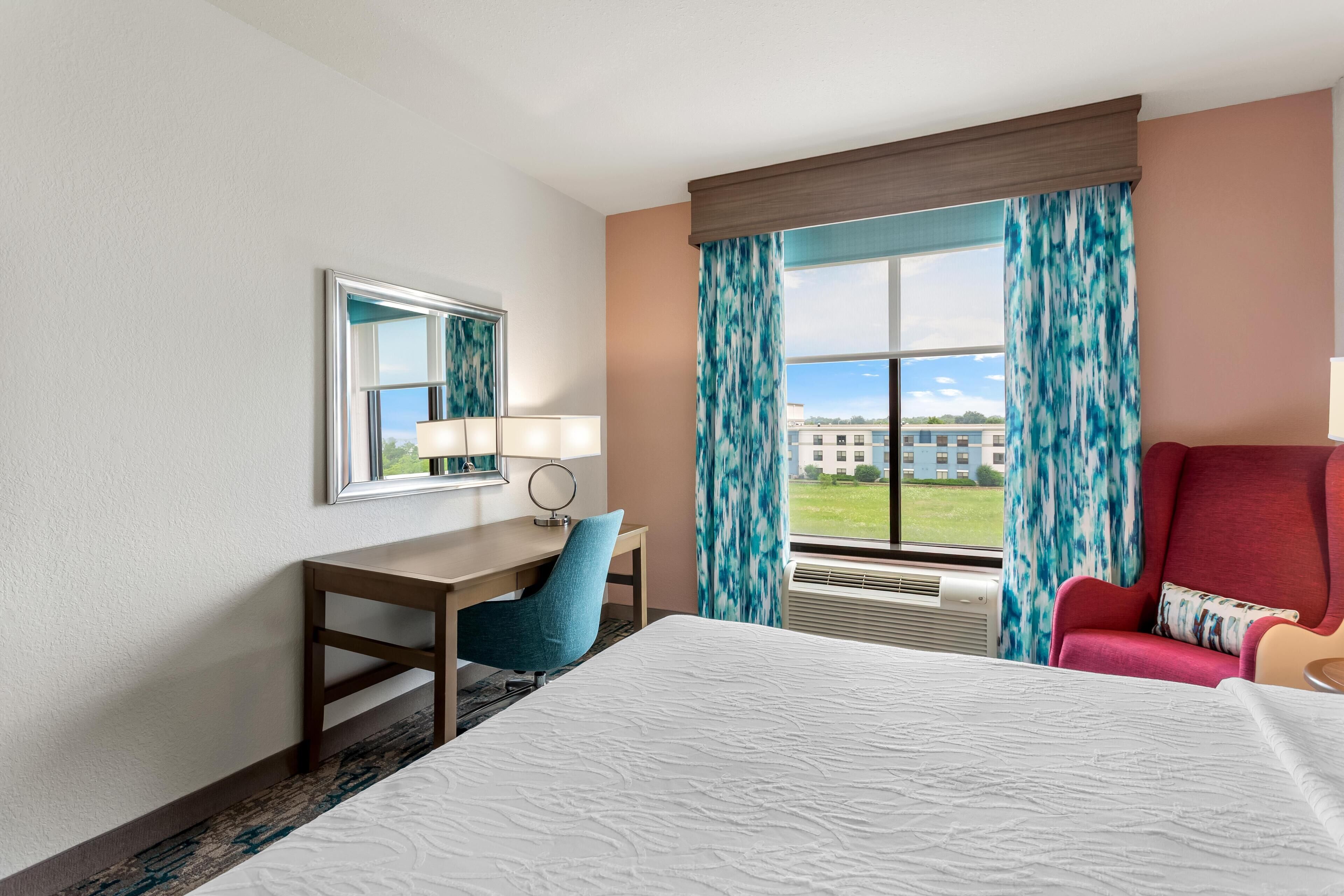 Habitación de hotel con cama, escritorio, silla, ventana con cortinas y vista a edificios.