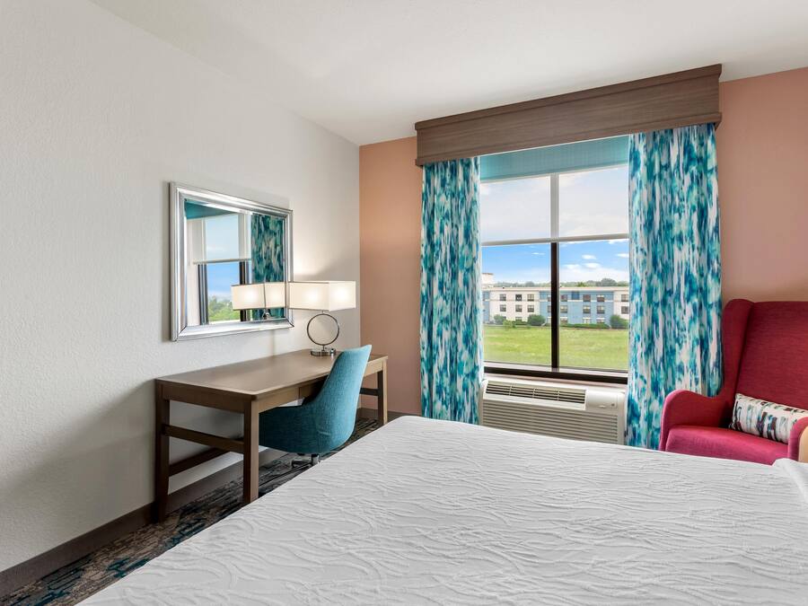 A hotel room with a bed, a desk, a chair, a window with curtains, and a view of buildings outside.