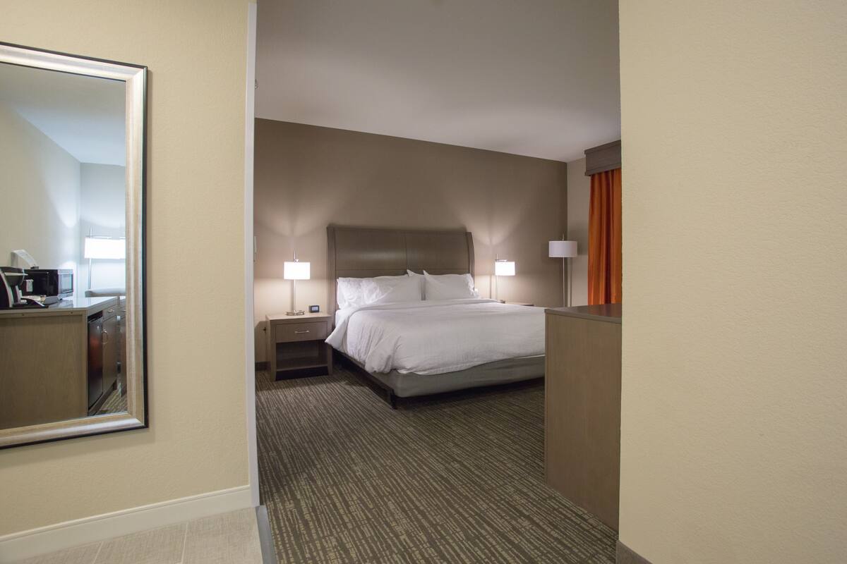 A hotel room with a large bed, two bedside tables with lamps, a mirror, and a view of a kitchen area.
