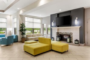 A spacious living room with a sectional sofa, a flat-screen TV mounted above a fireplace, and large windows offering a view of the outdoors.