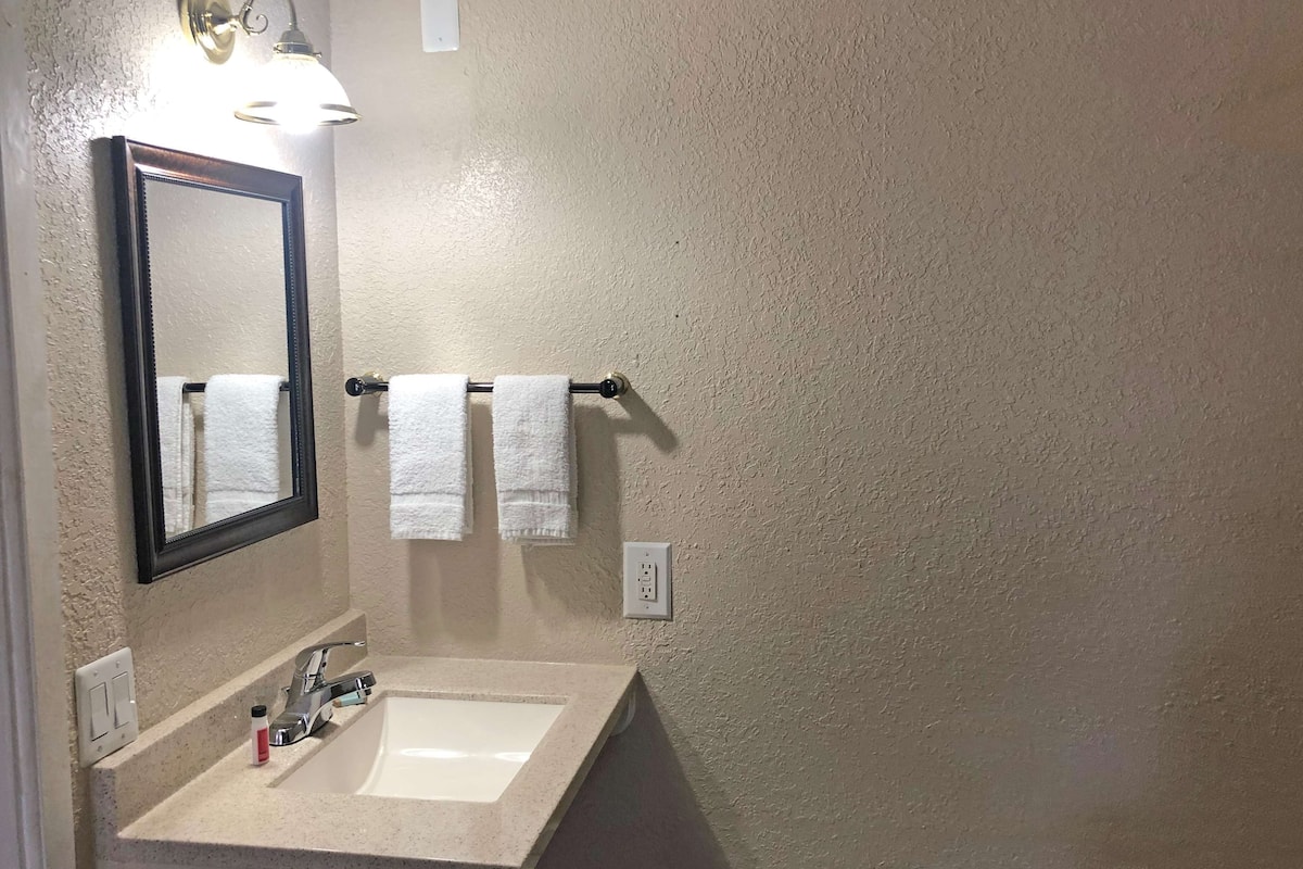 Un baño con un lavamanos blanco, un espejo y dos toallas blancas colgadas en un perchero.