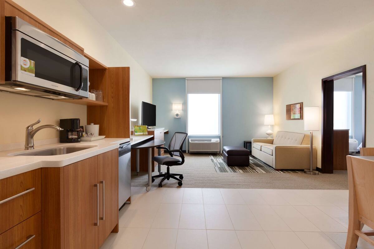 A modern kitchen with a microwave, sink, and coffee maker. There is a dining area with a table and chairs, and a living room with a sofa and a TV.