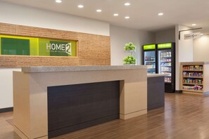 A reception area with a reception desk, a sign reading "HOME 2 SUITES BY HILTON", a refrigerator with a "COLD" sign, and a shelf with snacks. - Home2 Suites by Hilton Oklahoma City Quail Springs (Oklahoma City)