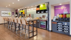 Un espace de restauration moderne dans un hôtel, avec une longue table conviviale, des tabourets de bar et un coin café équipé de récipients étiquetés.