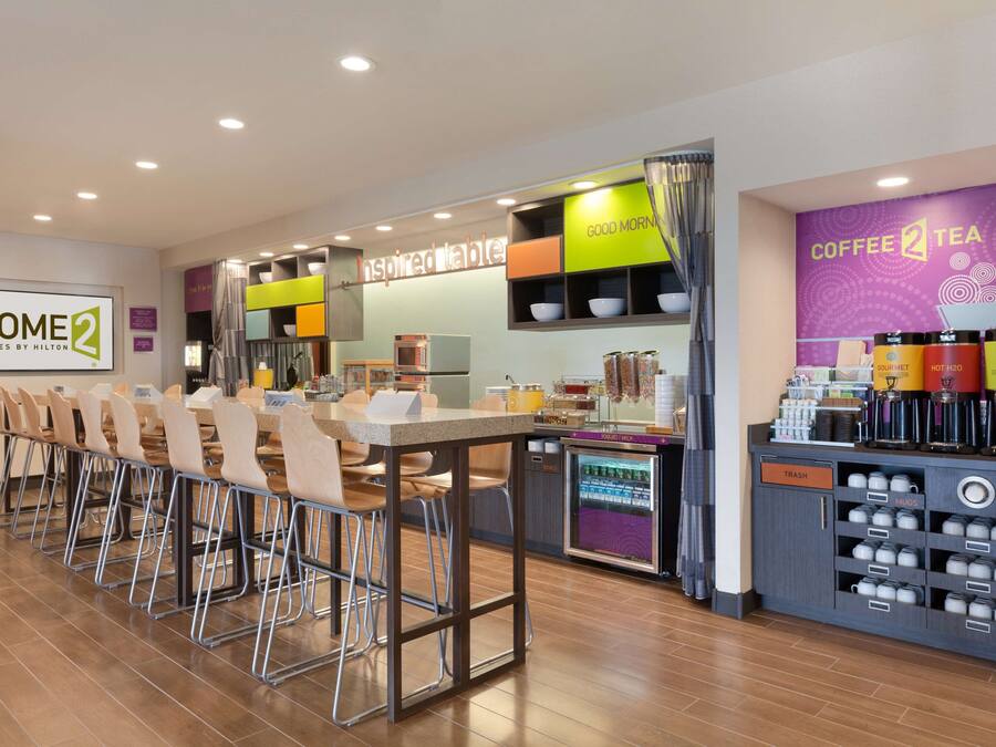 Uma área de refeições moderna em hotel, com uma mesa comunitária grande, banquetas de bar e um canto de café com recipientes identificados.