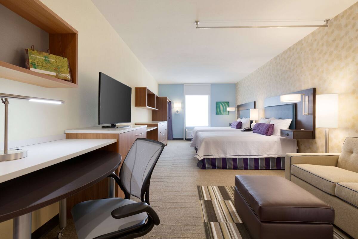 A hotel room with a desk, chair, television, two beds, a sofa, and a large window.
