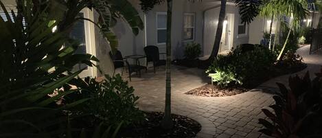 A paved courtyard with a seating area, surrounded by various plants and trees.