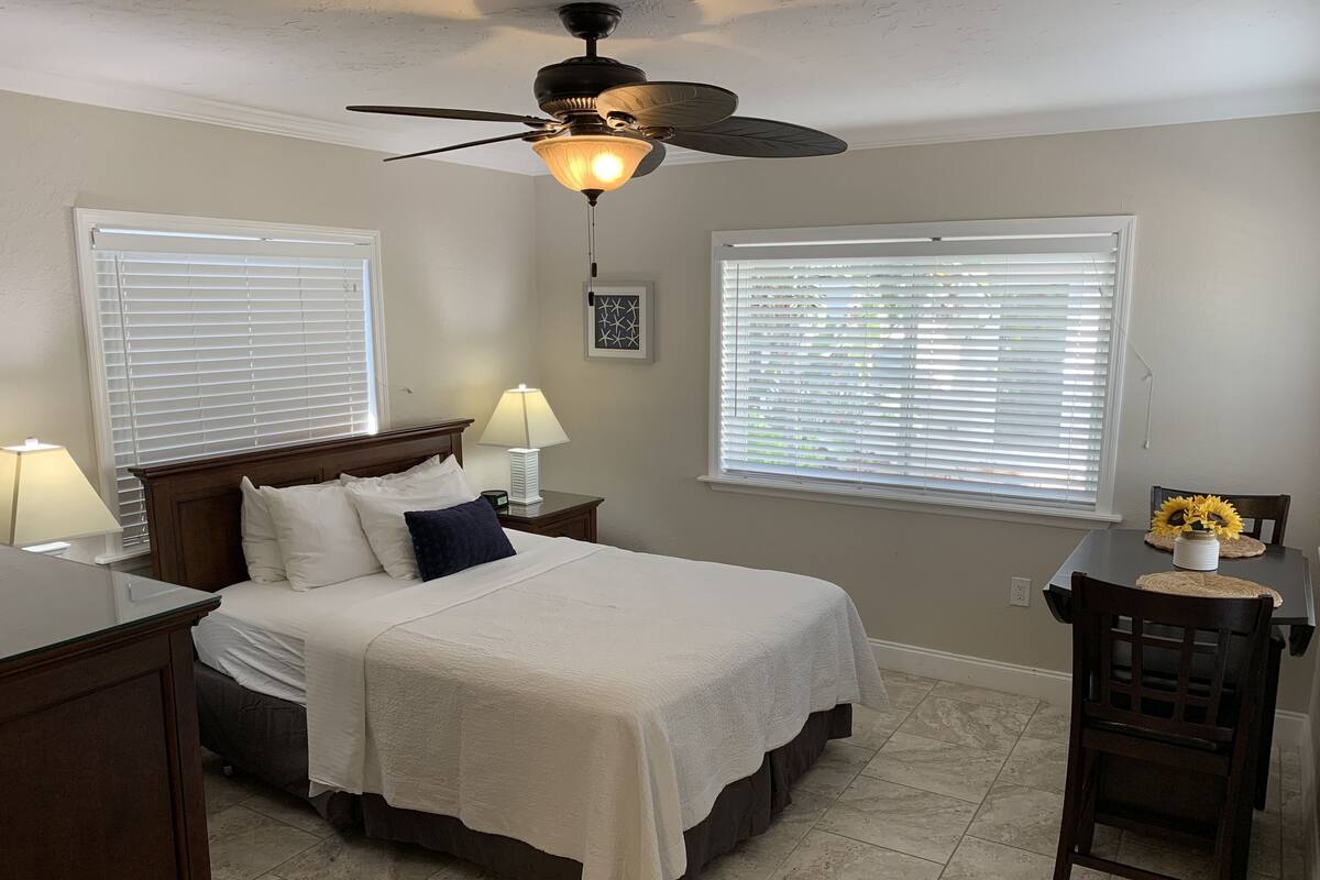 A bedroom with a bed, bedside lamps, a ceiling fan, and two windows with blinds.