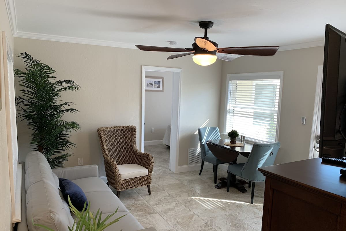 A living room with a beige sofa, a wicker chair, a dining table with chairs, a ceiling fan, and a large plant.