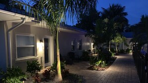Ein Wohngebiet mit einem gepflasterten Weg, einem Haus mit der Hausnummer 8 und einem Garten mit verschiedenen Pflanzen und einer Palme.