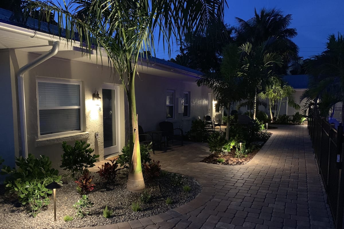 A residential area with a paved walkway, a house with a visible number '8' on the door, and a garden with various plants and a palm tree.