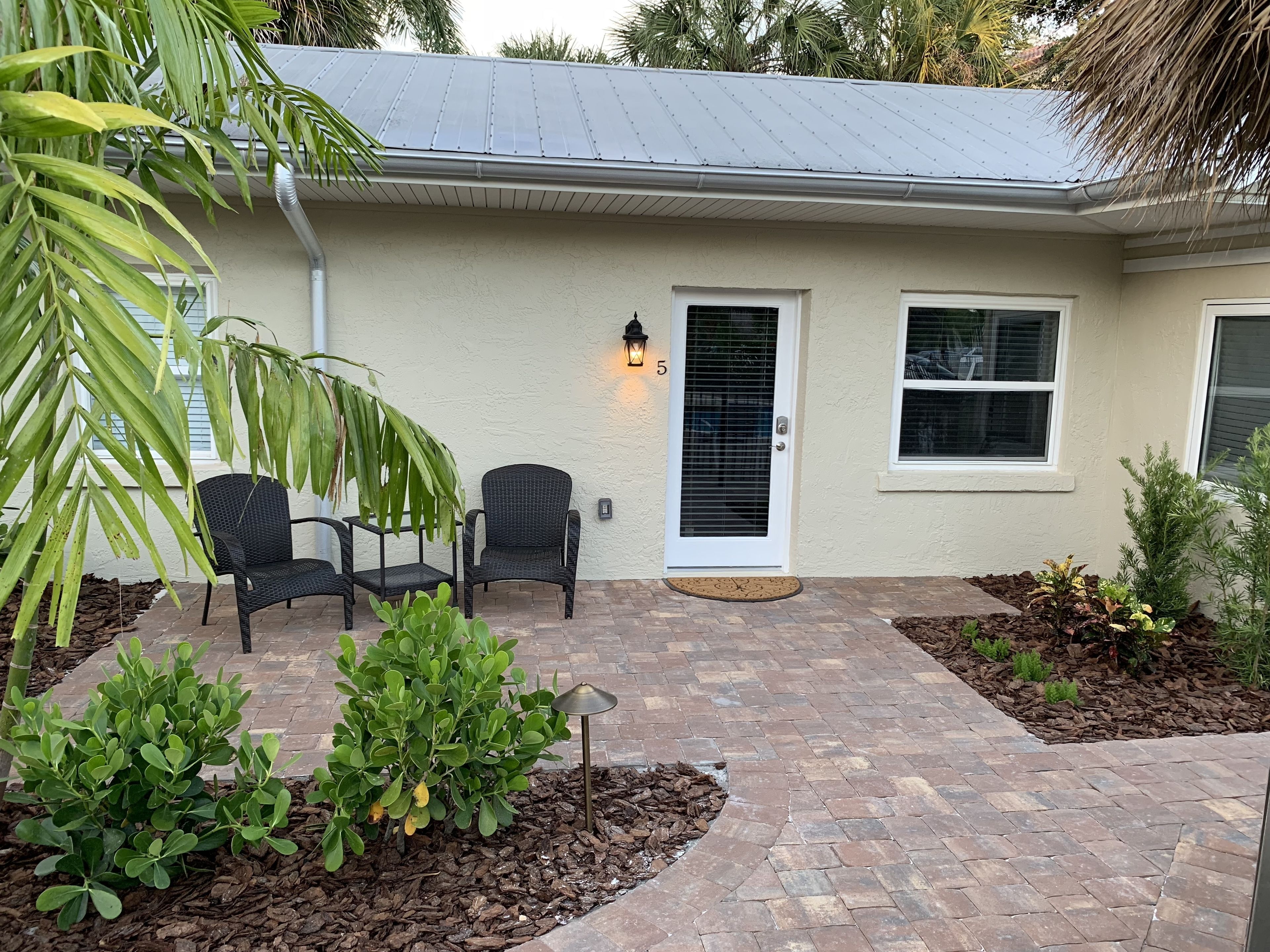 A house with a patio area featuring two wicker chairs, a small garden with mulch, and a metal roof.