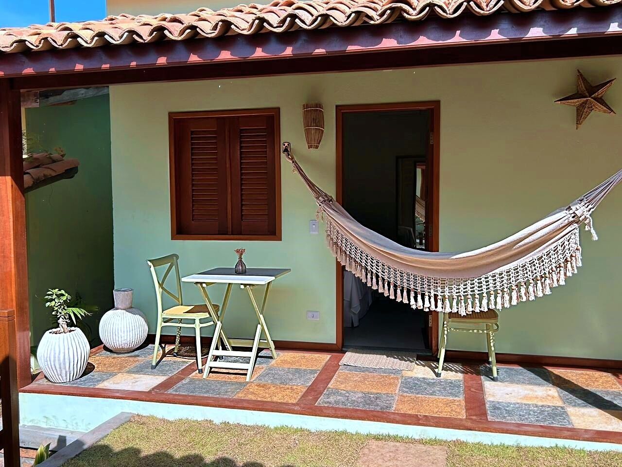 A small, single-story building with a tiled roof and a hammock. There is a small table and two chairs outside, and a star decoration on the wall.