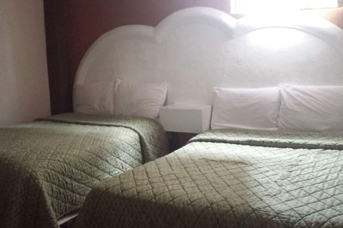 Une chambre avec deux lits simples, chacun recouvert d’une courtepointe matelassée verte et agrémenté d’oreillers blancs. Une table de chevet avec une lampe se trouve sur un des côtés.