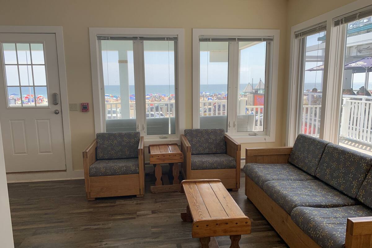 A room with a wooden floor, a sofa, two armchairs, a wooden coffee table, and large windows with a view of the beach and buildings.