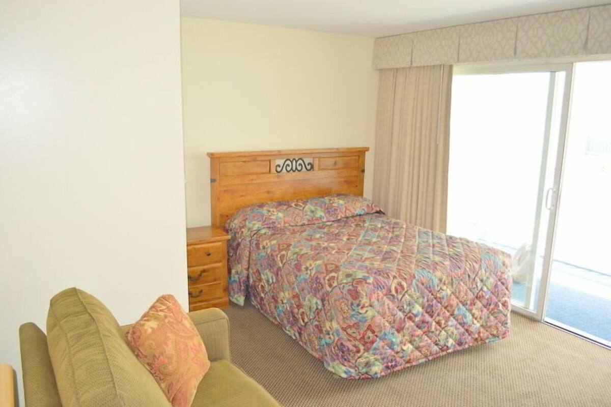 A hotel room with a wooden headboard, a colorful bedspread, a green armchair, and a sliding glass door to the outside.